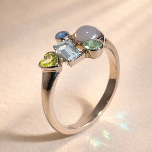 Ruinette Constellation Ring 1 shown at angle, displays an 18k white gold band with a cluster of five gemstones in Sapphire, Aquamarine, Gray Moonstone, and Peridot.
